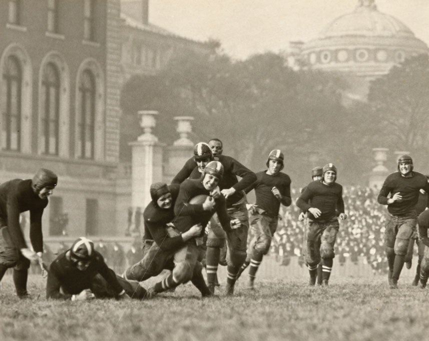 A man holding a football while being tackled, with teammates running towards him.