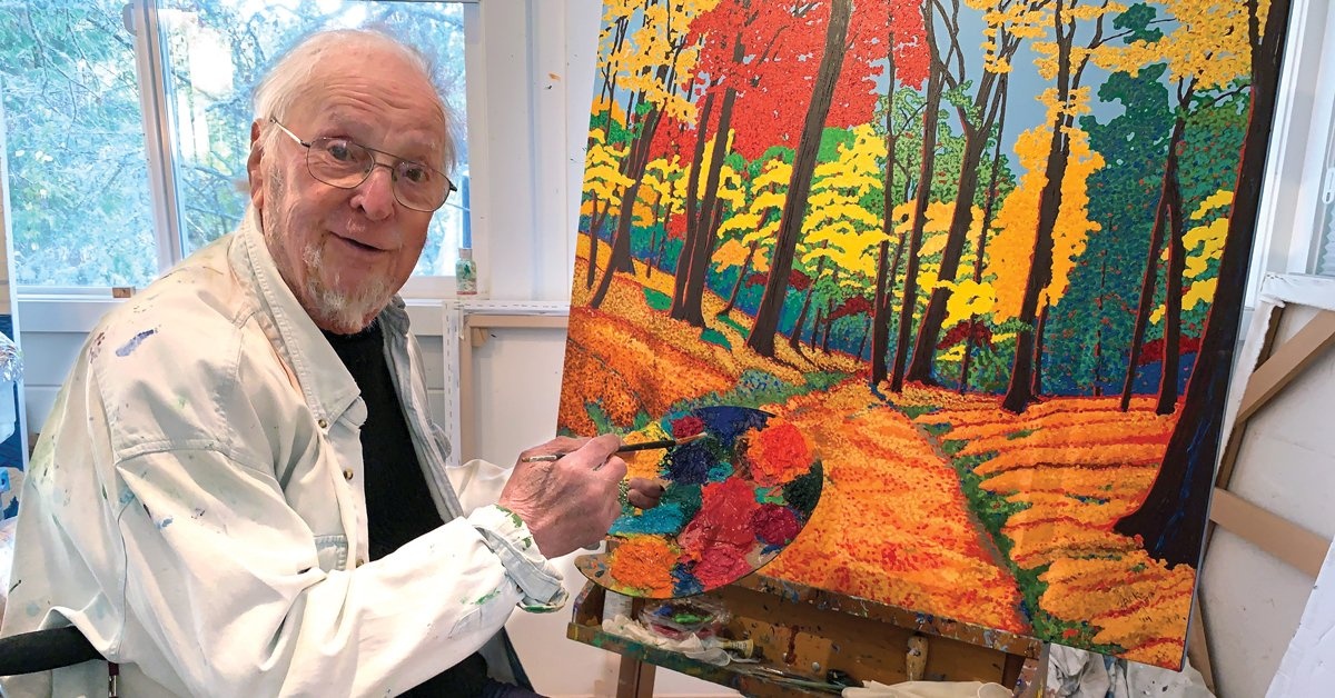 Jack Stuppin ’55 seated in front of an unfinished painting, holding a paintbrush