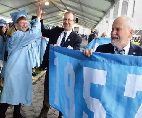 a graduate in cap and gown high-fiving a man in a suit with an older man smiling in the foreground