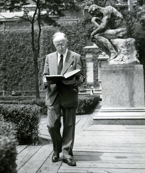 A man wearing glasses, reading a large book while walking.