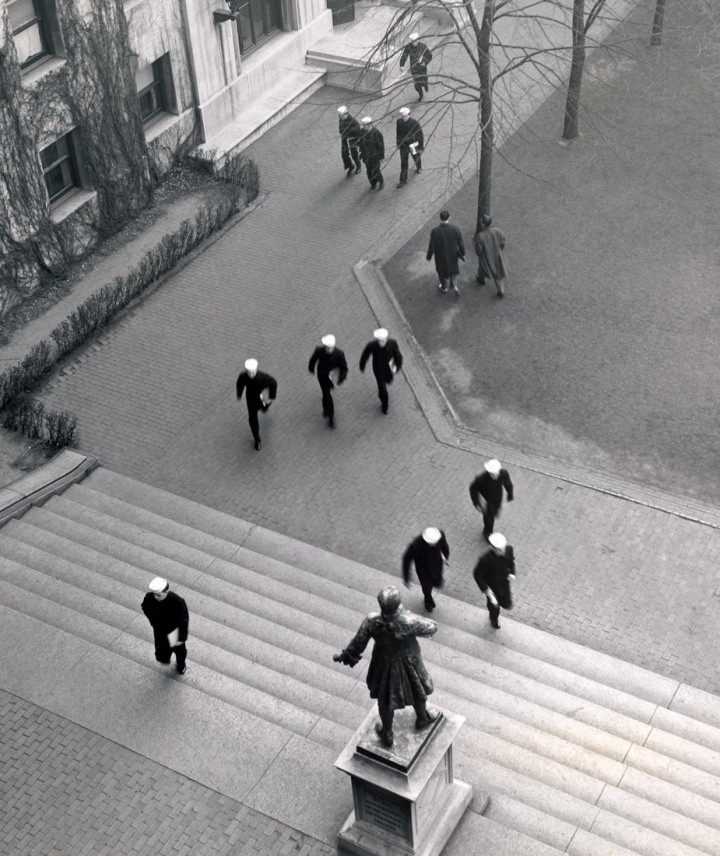 Several men walking on the Columbia University campus, a number of whom are  wearing dark uniforms and light-colored hats.