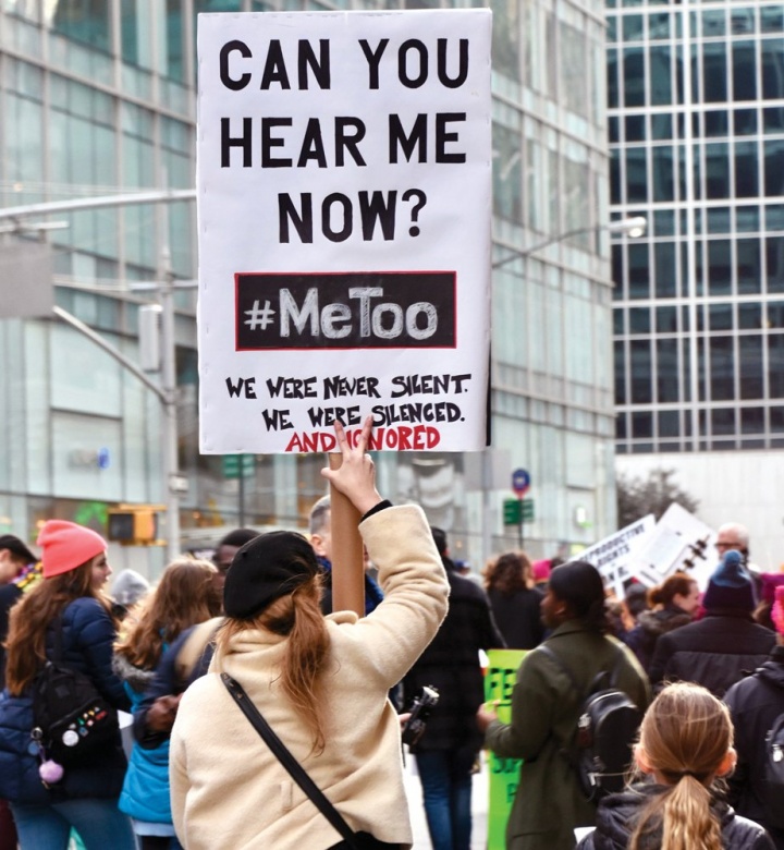 Several people outdoors in front of tall buildings holding signs, one of which contains the words "Can you hear me now? #MeToo."