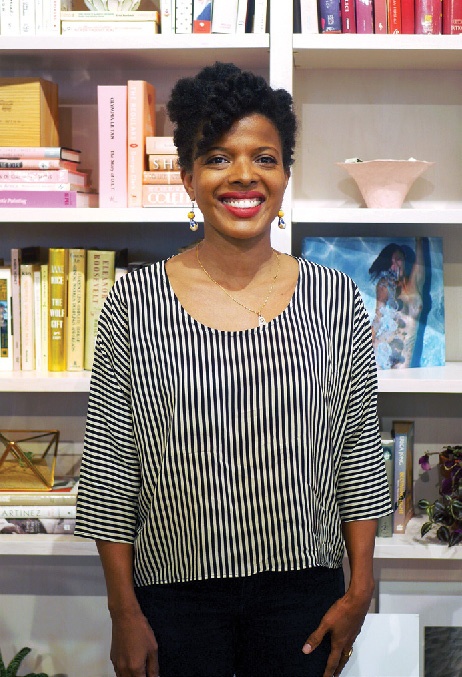 A black woman wearing red lipstick and smiling in front of a bookshelf