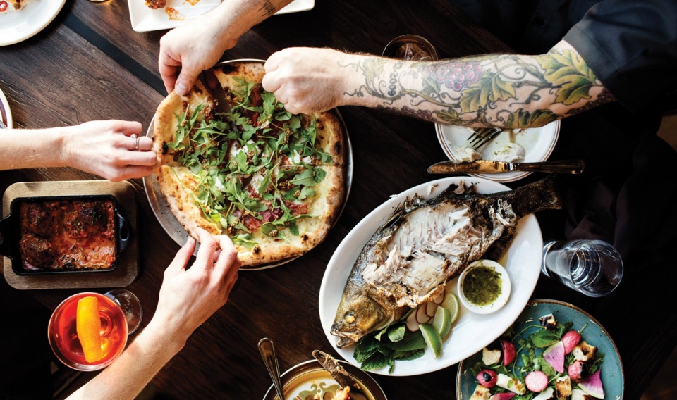 The hands and arms of several people reaching for portions of a pizza pie on a wooden table.