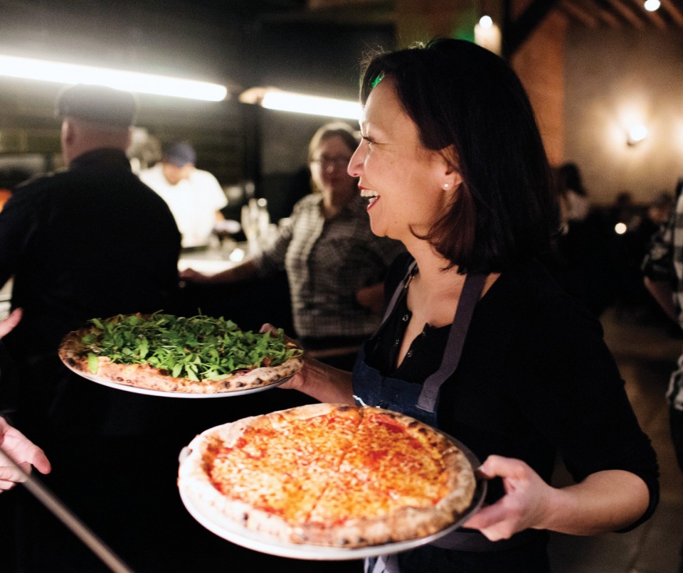 A woman smiling holding two pizzas in dishes