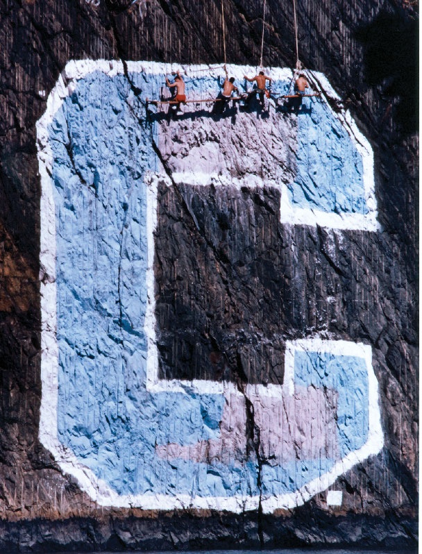 Four rowers apply paint to a large "C" insignia located on a cliff.