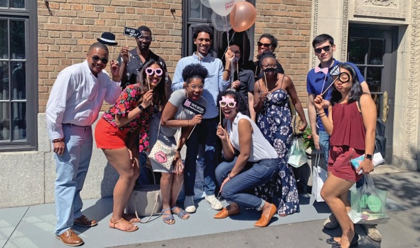 Several people smiling, outside of a building on a sunny day, one of whom is holding balloons